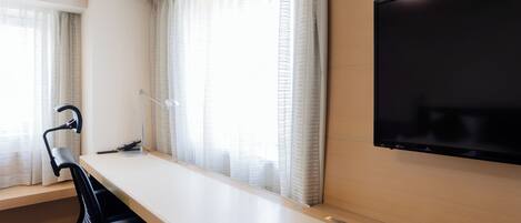 A modern hotel room with a large desk, a flat-screen TV mounted on the wall, and large windows with sheer curtains.