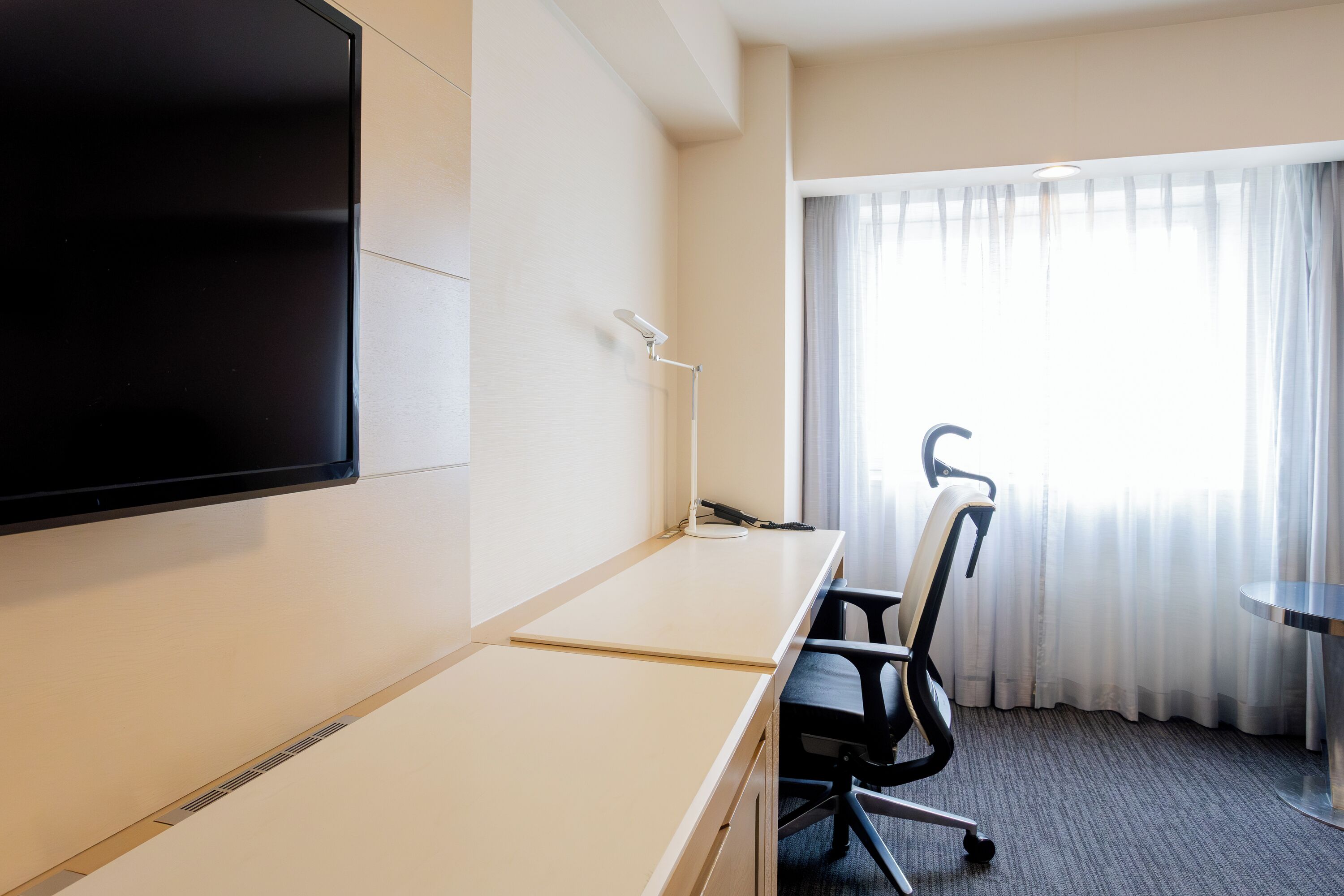 Habitación de hotel con televisión de pantalla plana en la pared, escritorio con silla, lámpara y ventana con cortinas.
