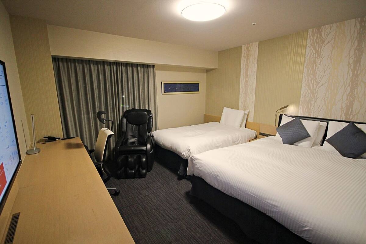 Een hotelkamer met twee bedden, een massagestoel, een bureau met een computer en een raam met gordijnen.