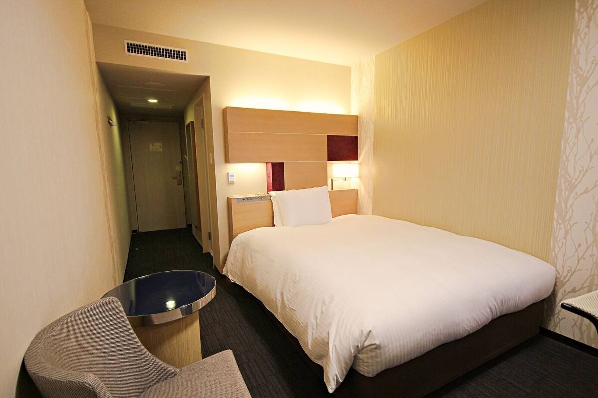 Een hotelkamer met een bed, een kleintje tafeltje en een stoel. Er is een deur naar een andere kamer en een ventilatie rooster aan het plafond.