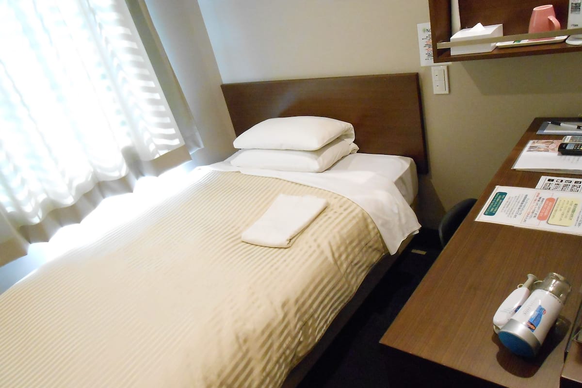 A hotel room with a bed, a bedside table with a bottle and a tissue box, and a window with curtains.