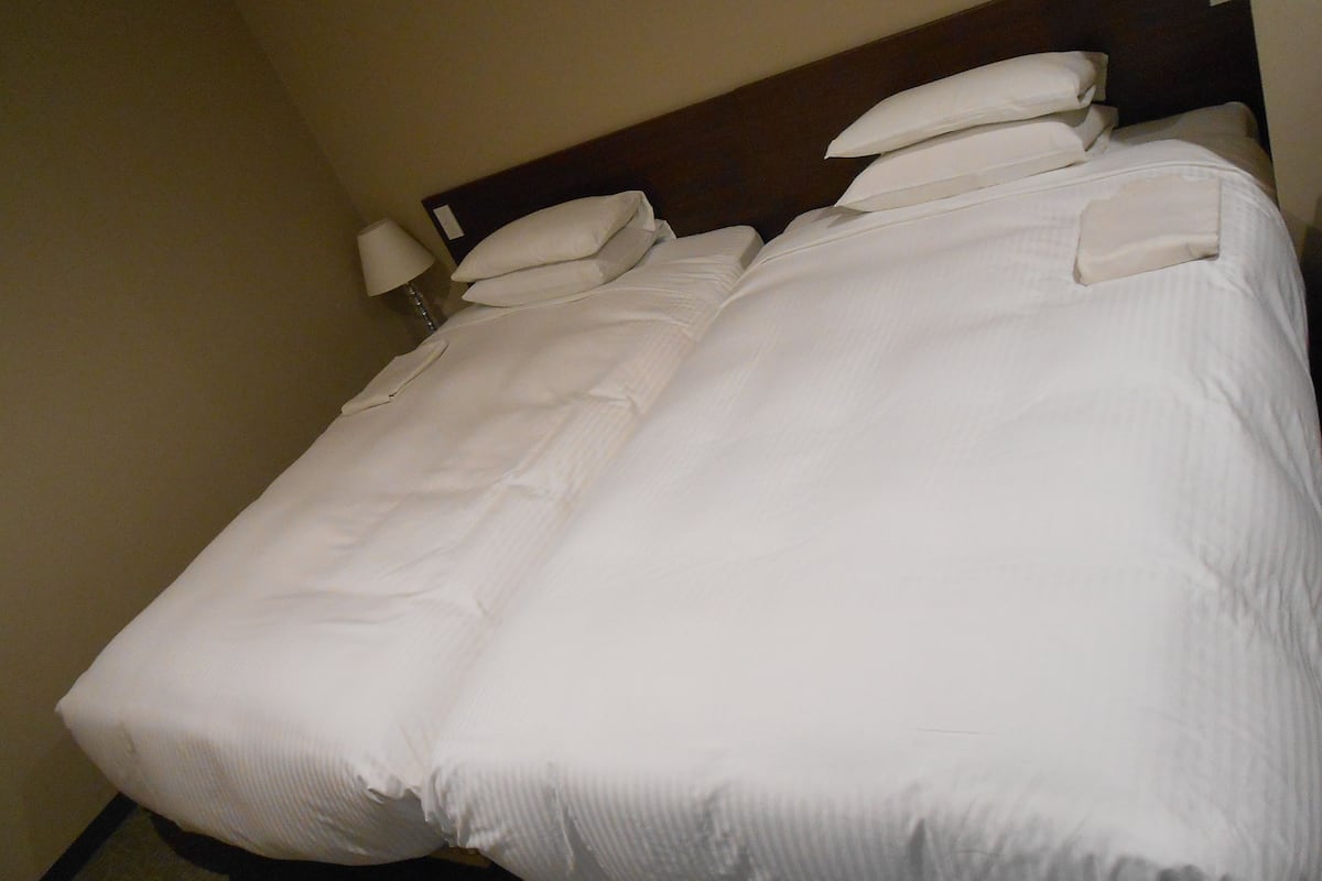 A neatly made bed with white linens and pillows, a wooden headboard, and a bedside lamp.