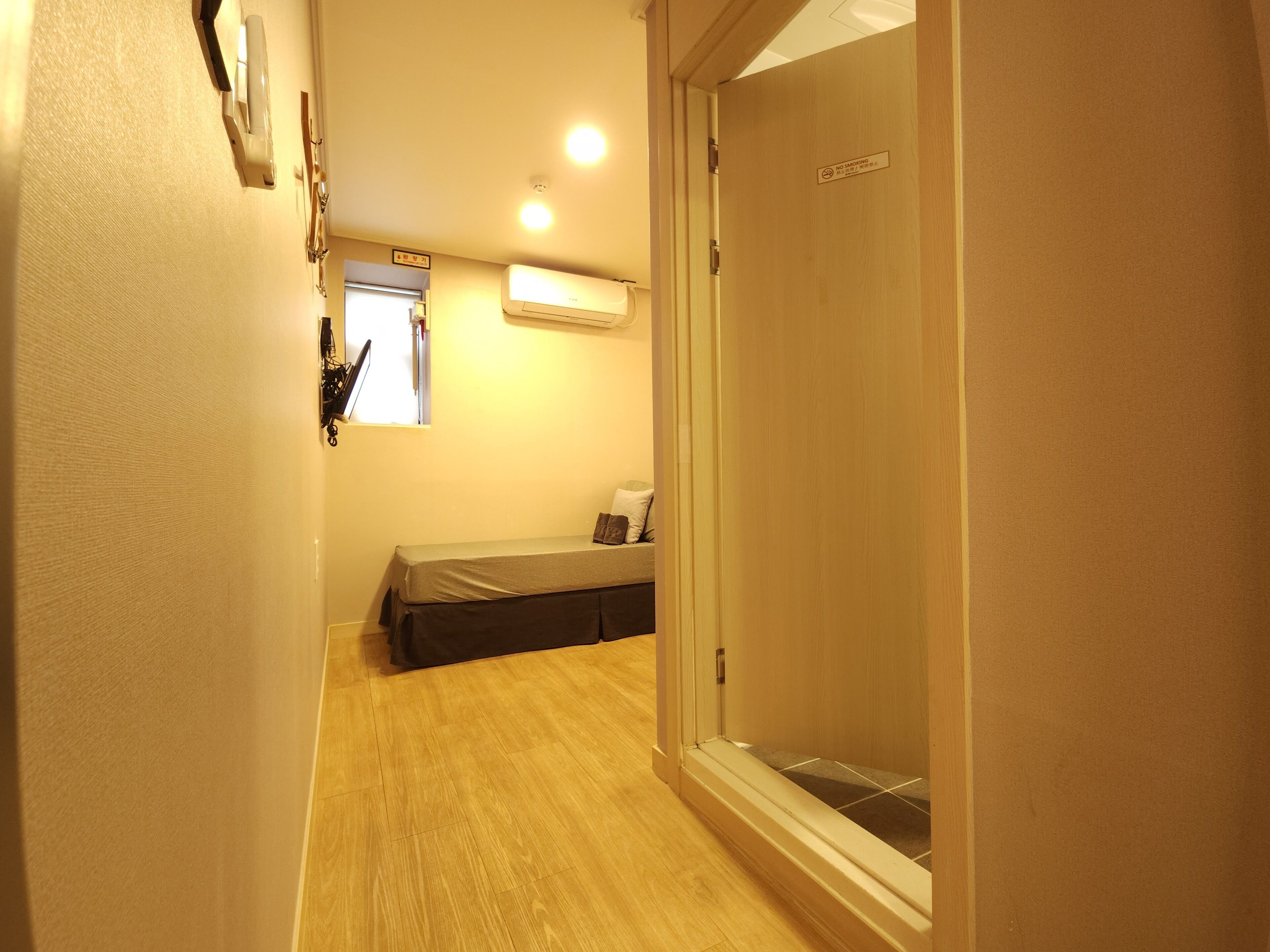 A narrow hallway with a wooden floor leading to a room with a bed, a window, and a wall-mounted air conditioner.