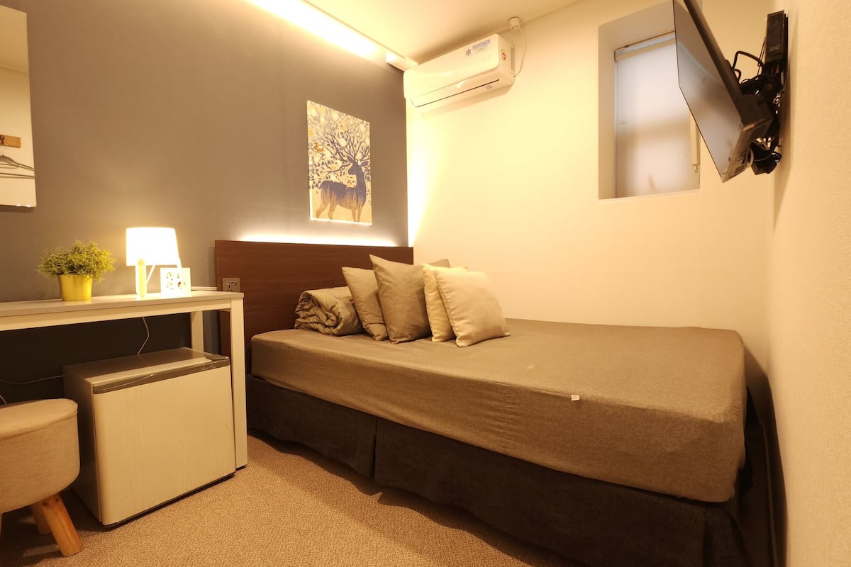 A compact bedroom with a bed, bedside table, lamp, and a small desk with a chair.