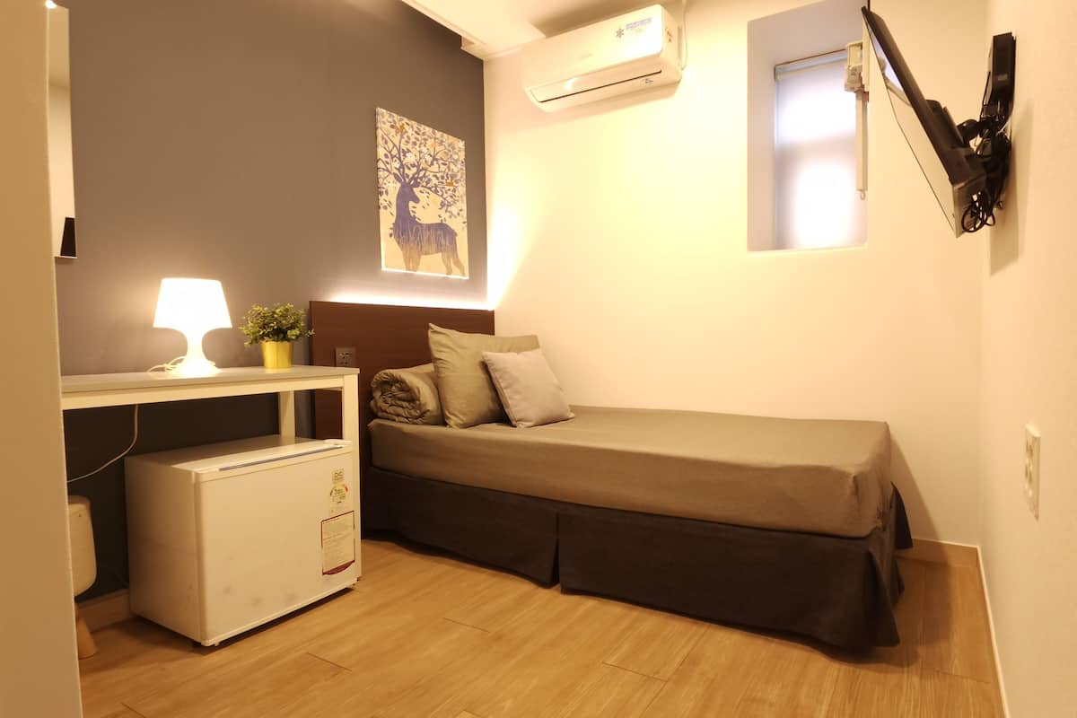 A small, compact room with a bed, a desk with a lamp, a small fridge, and an air conditioning unit.
