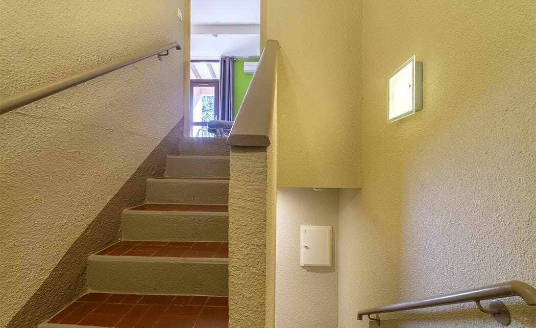 A staircase with white walls and a handrail, leading to a room with a green wall and a window.