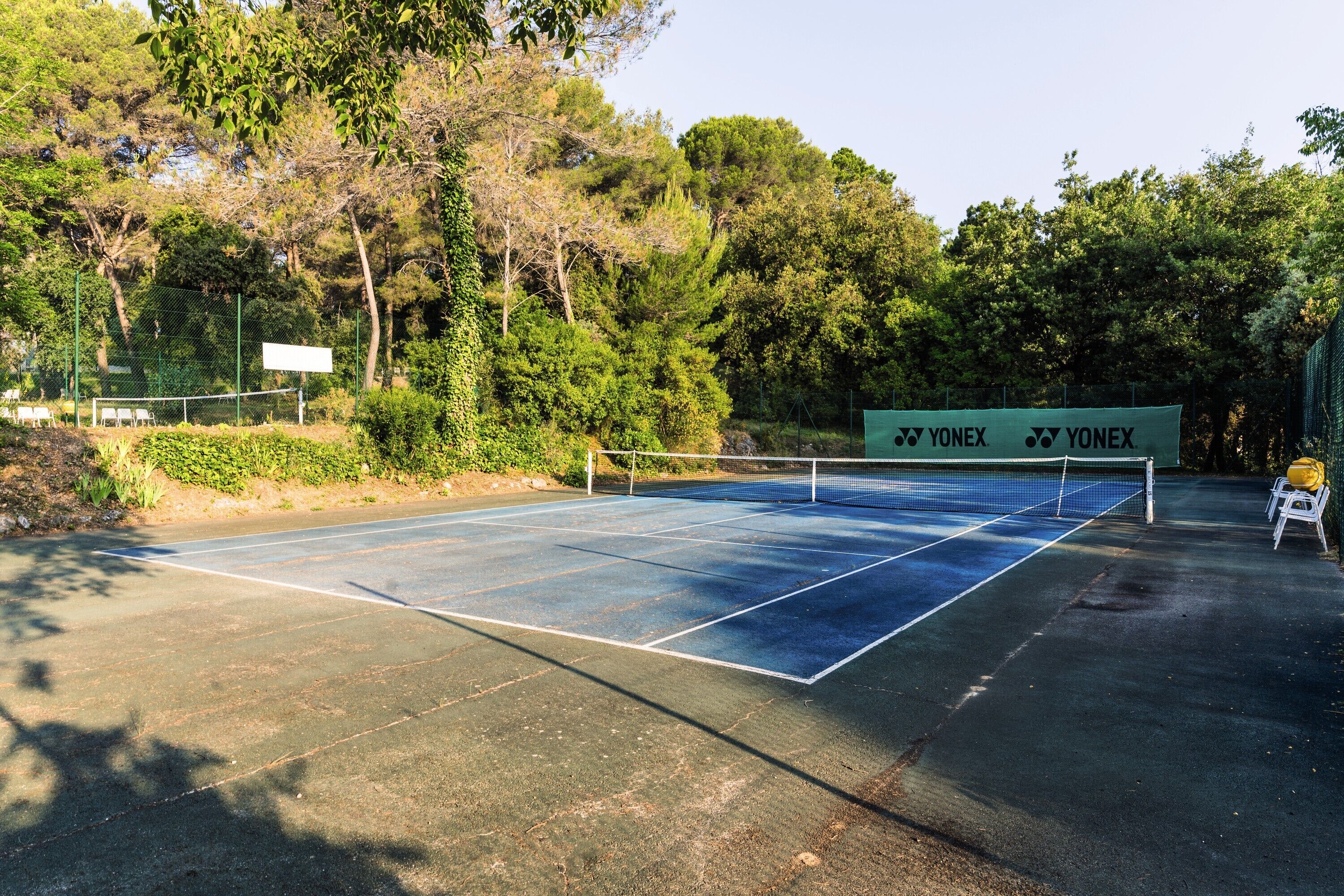 An outdoor tennis court with a Yonex brand sign, surrounded by trees and greenery.