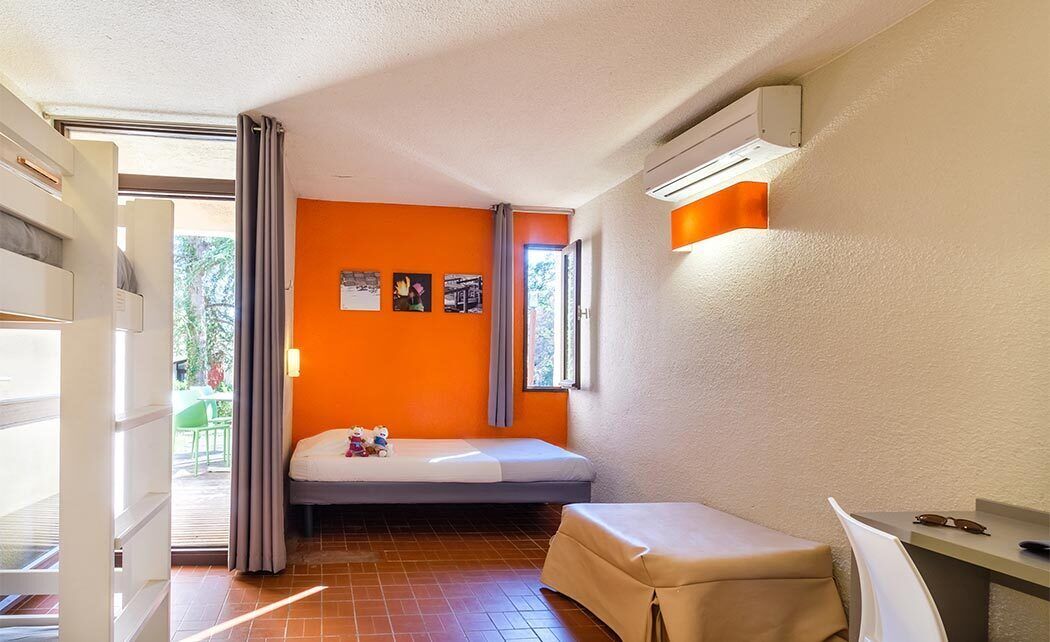 A compact room with a bed, a small table, and a chair. There is a window with curtains, a door, and a wall-mounted air conditioning unit.