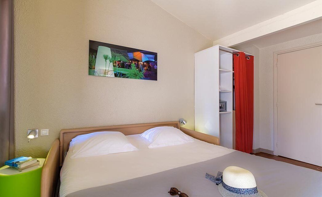 Een hotelkamer met een bed, nachtkastje, een wandplank en een deur met rode gordijnen.