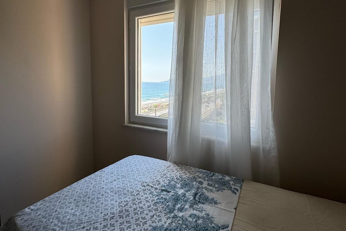 Une chambre à coucher avec un lit, une fenêtre ornée de rideaux légers, une vue sur la mer et un plafond doté d’un éclairage encastré.