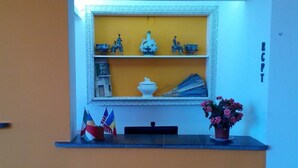 A reception area with a yellow wall, a framed shelf with decorative items, and a potted plant. - M'ama Holiday Rooms (Palermo)
