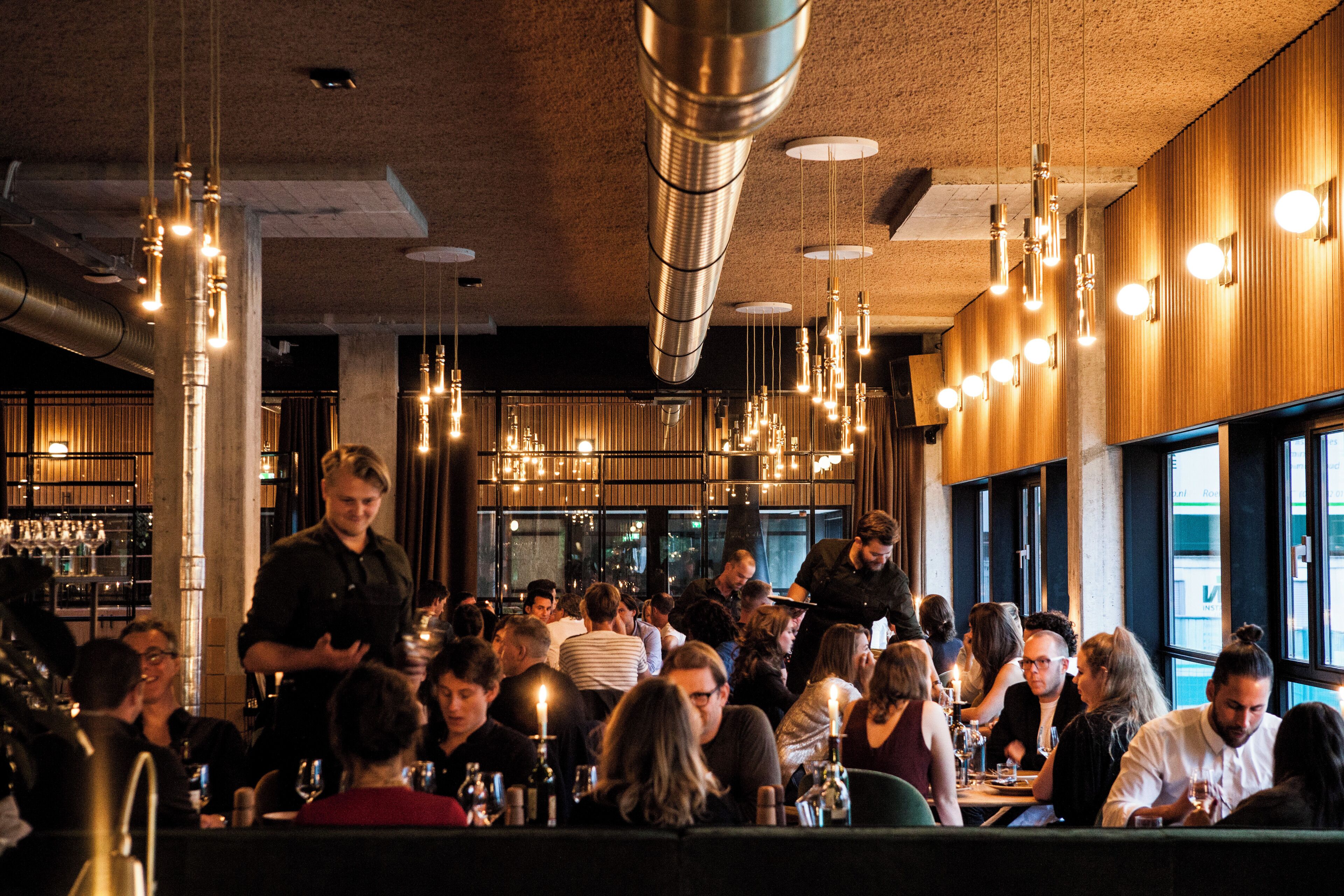 Restaurante movimentado com luminárias pendentes, um bar e clientes jantando.