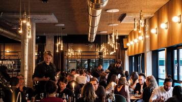 Restaurante movimentado com luminárias pendentes, um bar e clientes jantando.