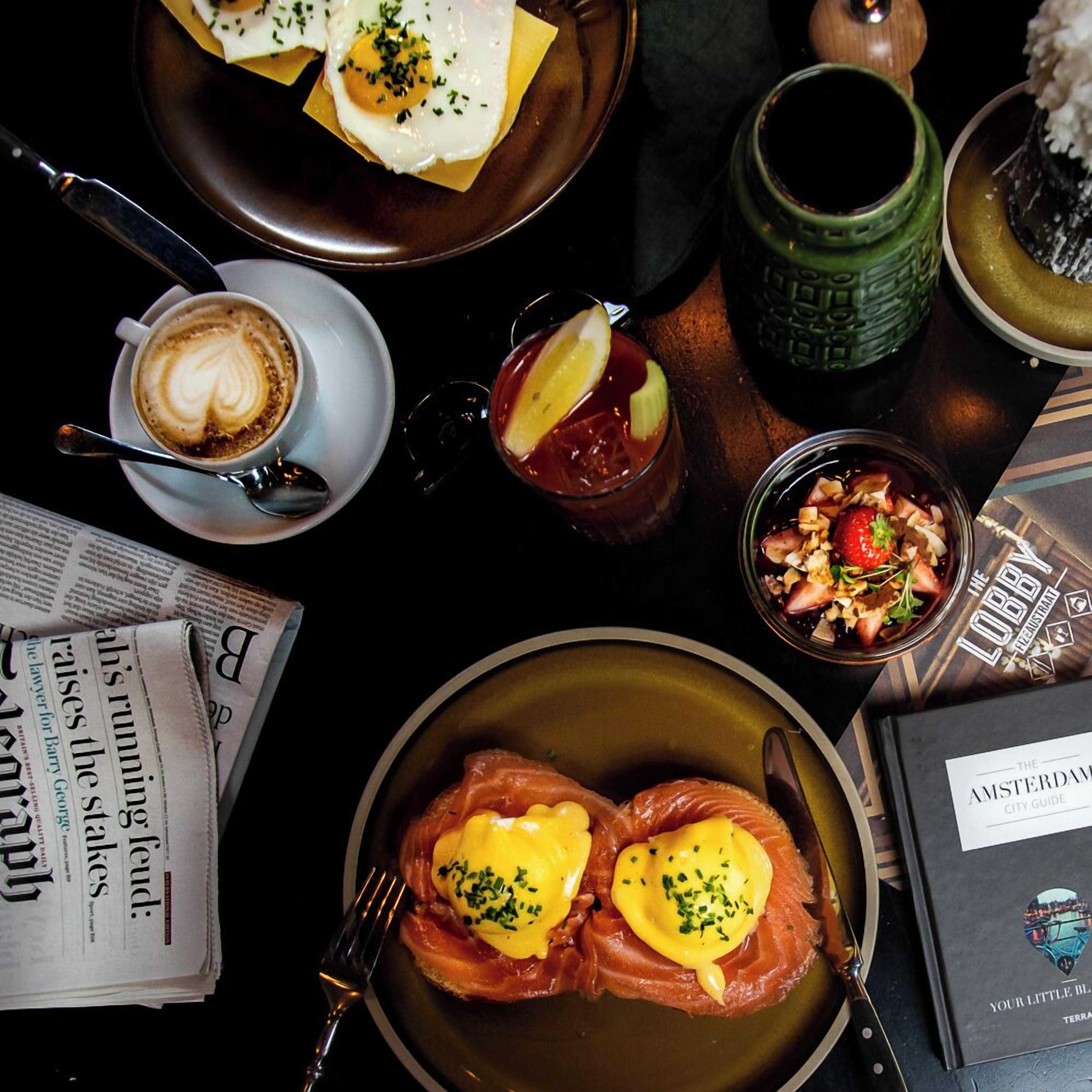 Una mesa con diferentes alimentos, una taza de café, un vaso de bebida roja, un libro y un periódico.