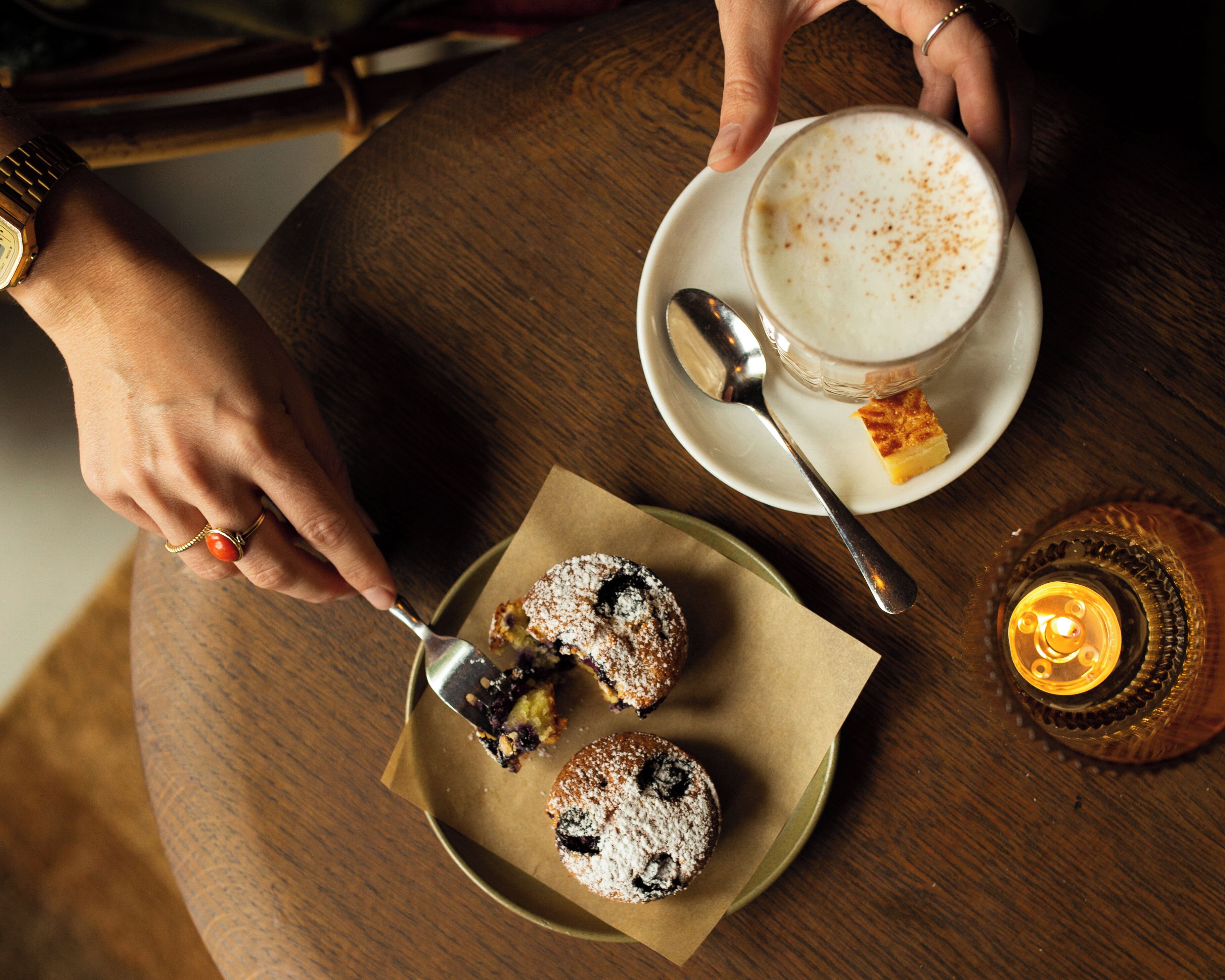 A person serving a dessert with a spoon, a cup of coffee on a saucer, and a candle on the table.