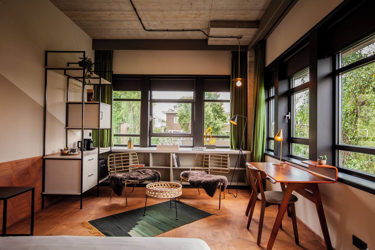 Une pièce moderne et bien éclairée, avec un bureau en bois, des chaises, une bibliothèque et de grandes fenêtres donnant sur un paysage verdoyant.
