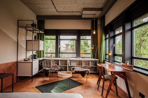 A modern room with a wooden desk, chairs, a small table, a bookshelf, and large windows.