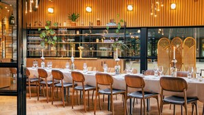 Un restaurante con paredes de paneles de madera, una larga mesa de comedor con sillas, y estanterías con cristalería y plantas.