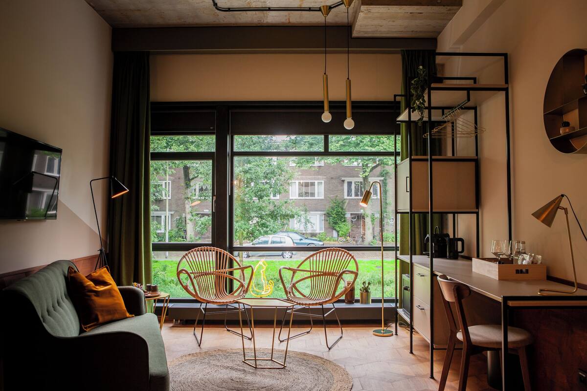 Un salon moderne comprenant un canapé, deux fauteuils, une table basse, un bureau et une grande fenêtre donnant sur un quartier résidentiel.