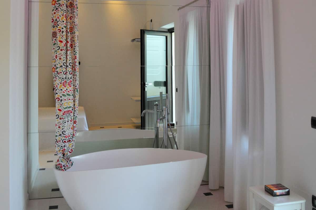 A modern bathroom with a large white bathtub, a shower with a colorful curtain, and a small white table with a book.