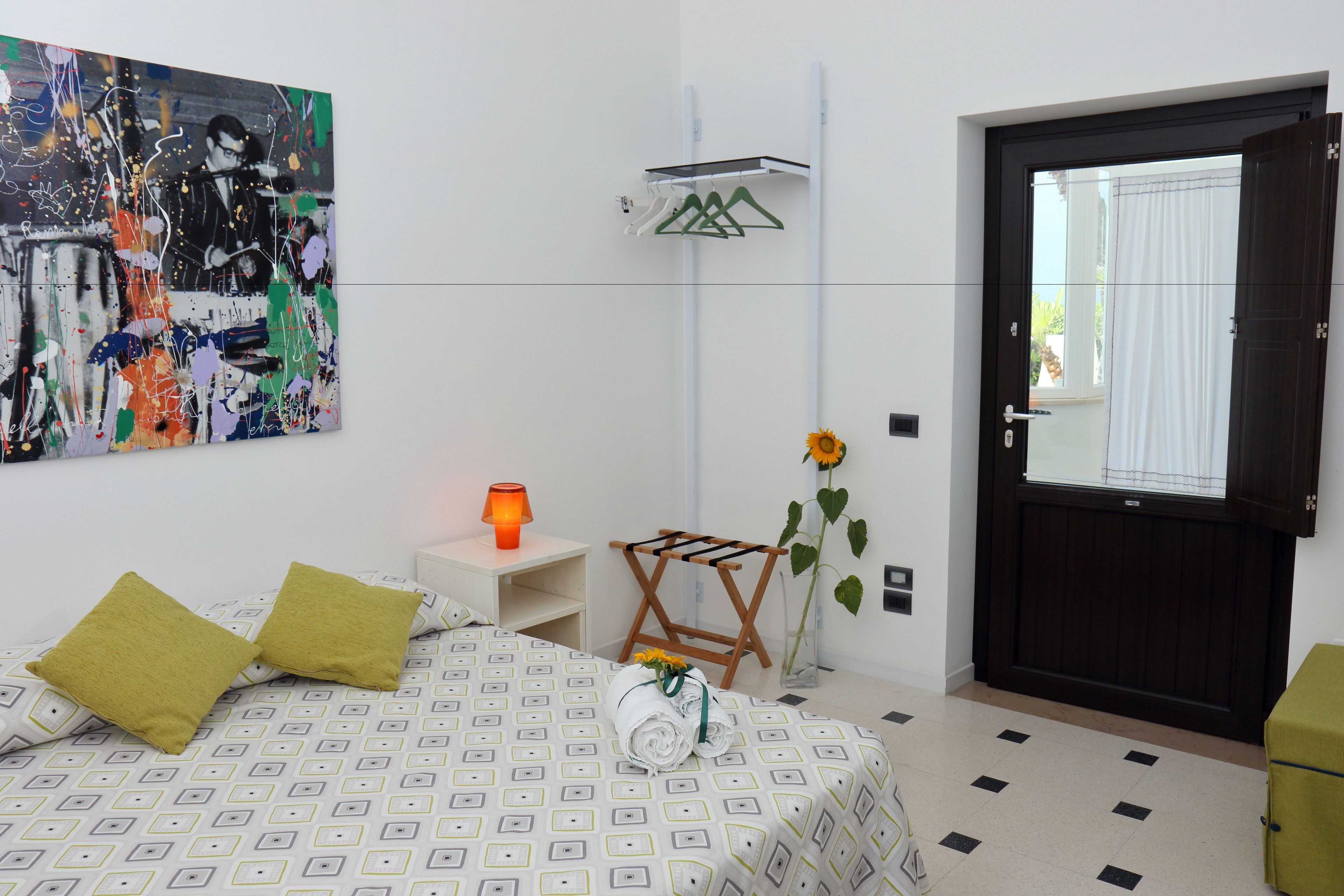 A hotel room with a bed, a painting, a nightstand, a lamp, a drying rack, a vase with a sunflower, and a door.
