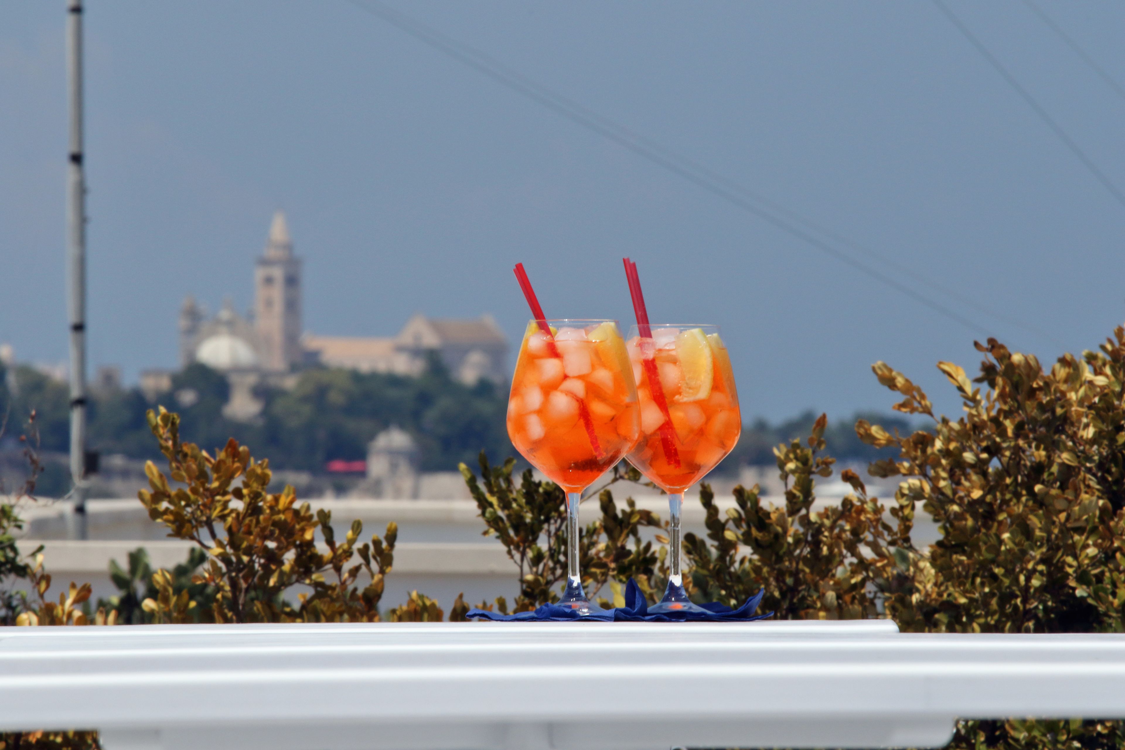 Dos vasos de bebida naranja con pajitas rojas sobre una superficie blanca, con un paisaje urbano y un edificio al fondo.