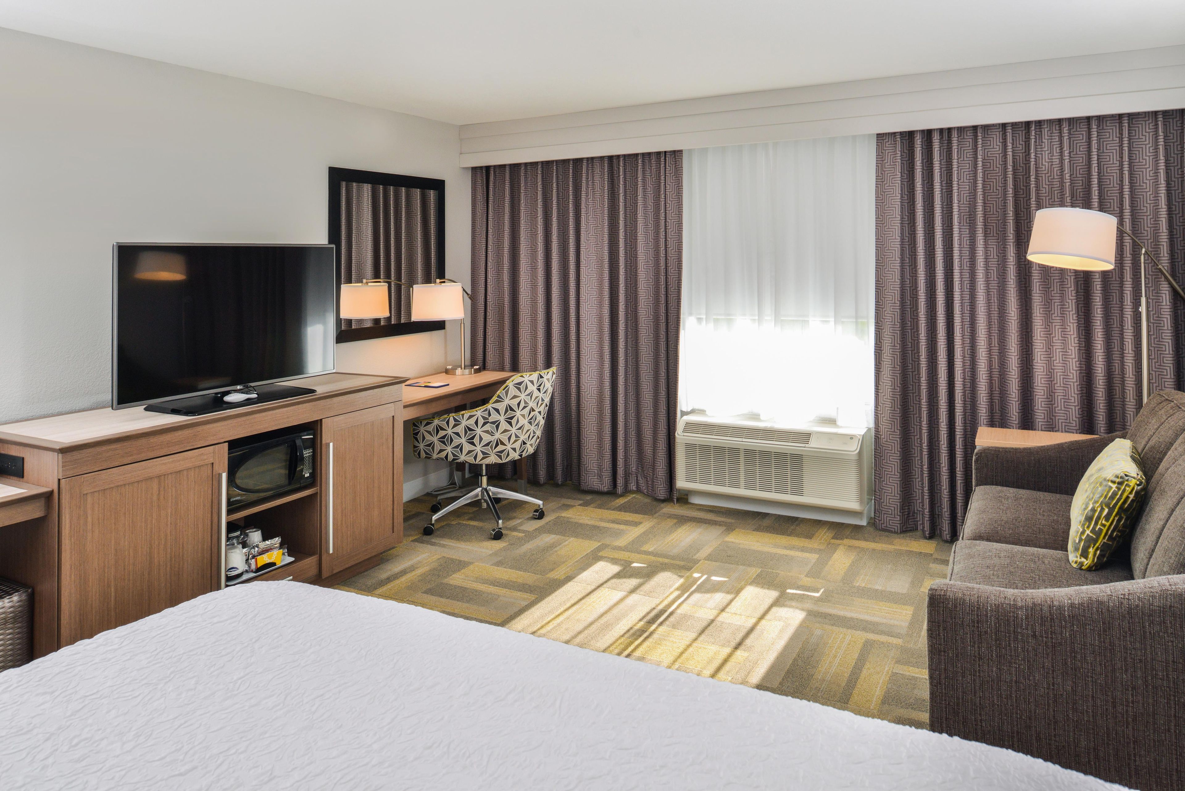 Une chambre d’hôtel comprenant un lit, un bureau avec un téléviseur à écran plat, une chaise, un canapé et une fenêtre avec des rideaux.