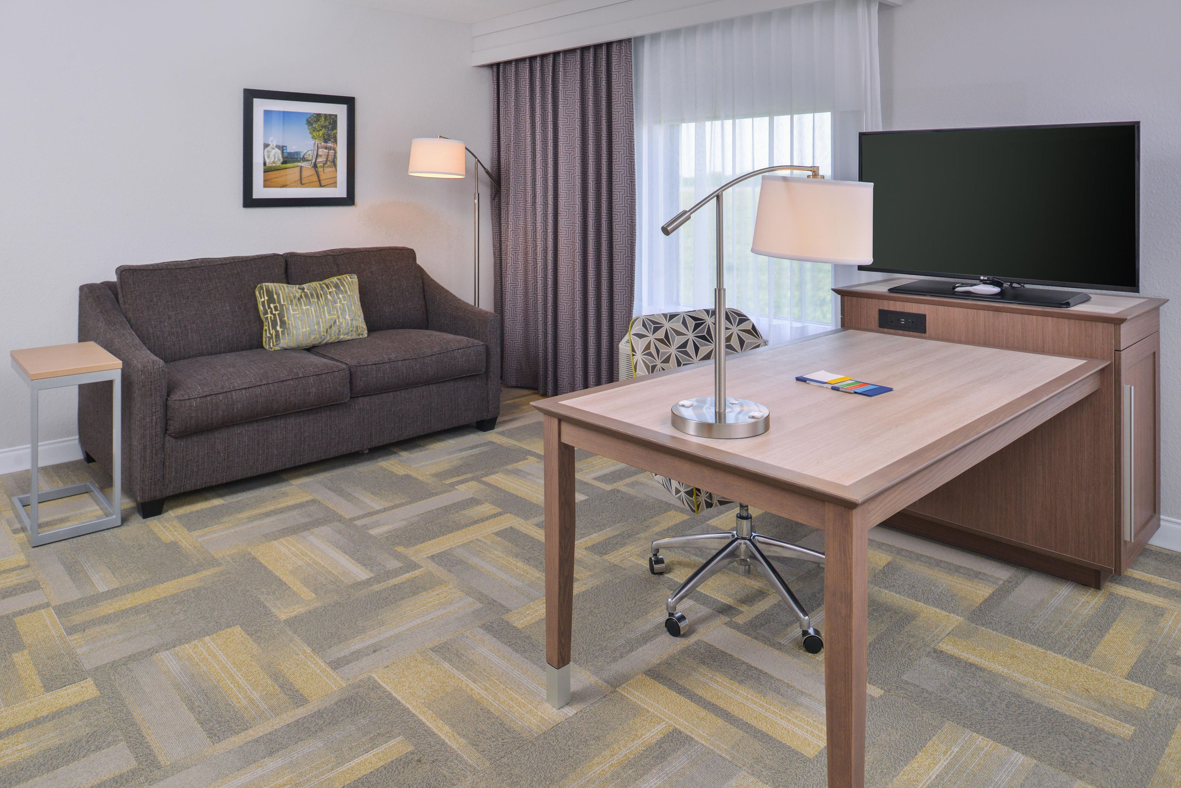 Habitación de hotel con sofá, escritorio con televisor, lámpara y alfombra estampada.