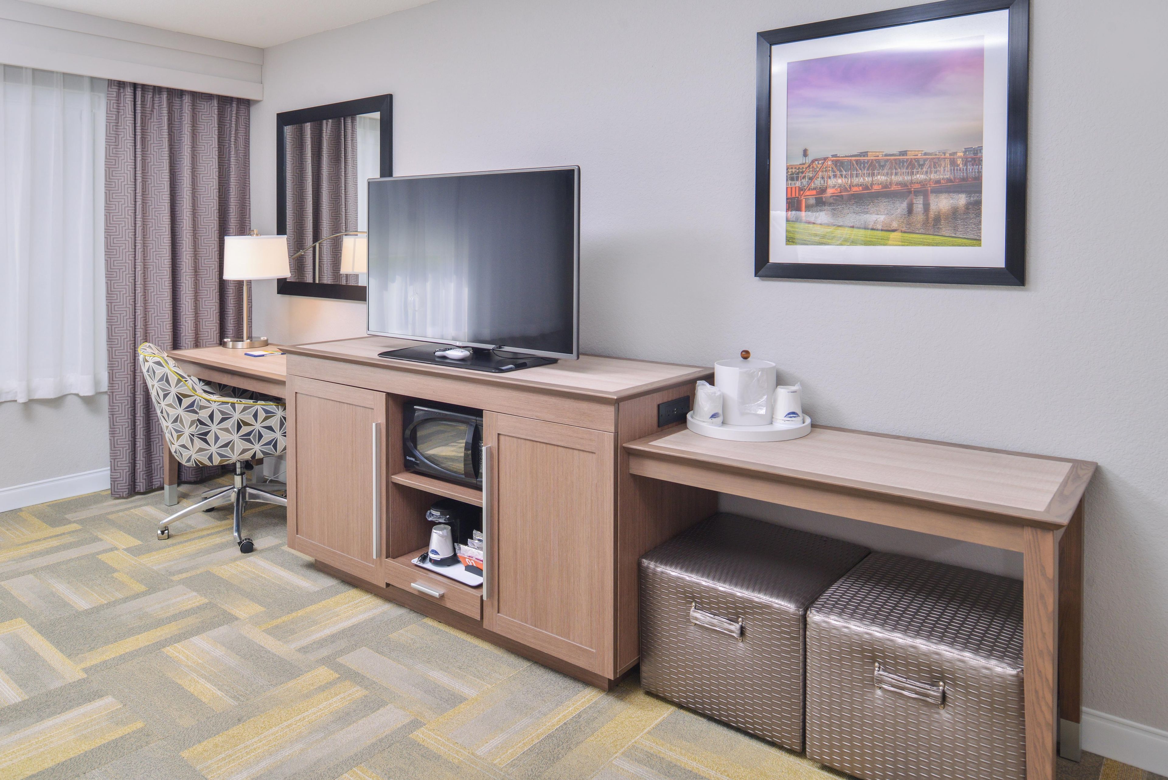 A hotel room with a flat-screen TV, a desk with a lamp, a chair, a small refrigerator, and a framed picture on the wall.