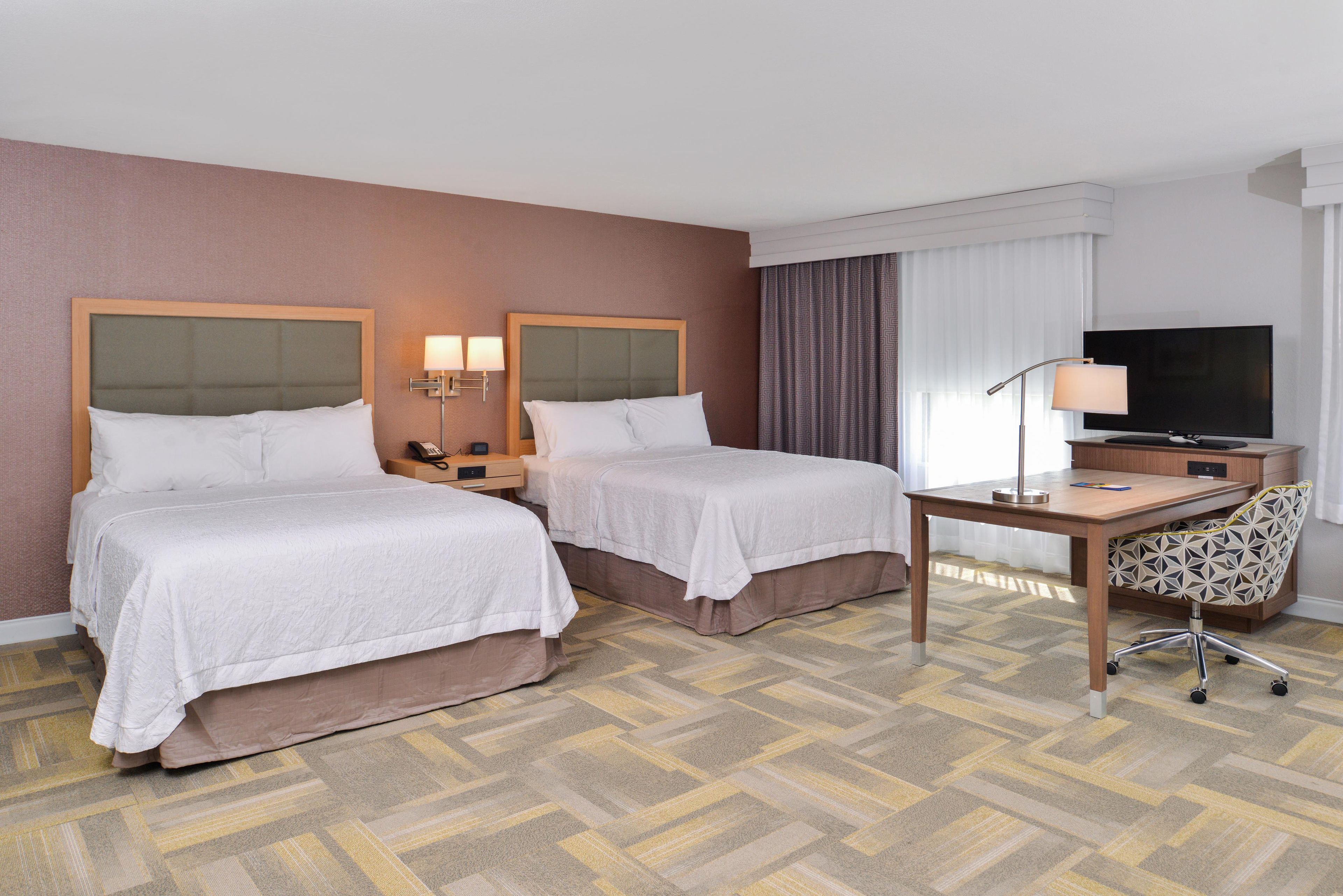 A hotel room with two beds, a desk, a chair, a television, and a window with curtains.