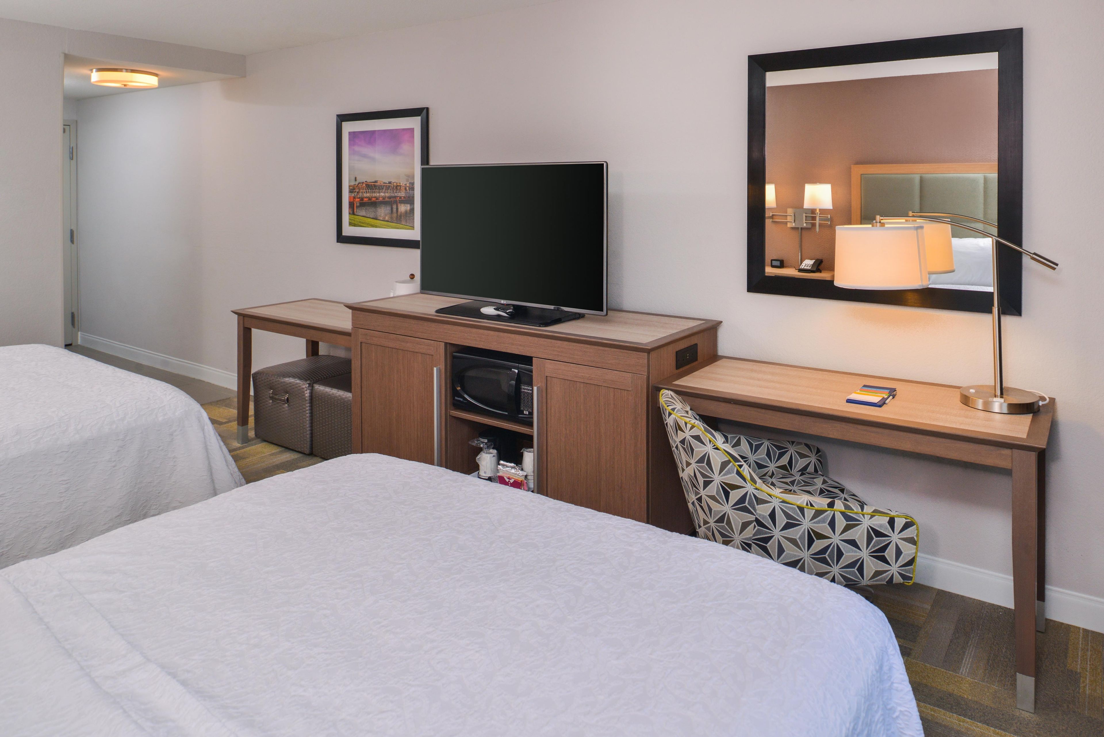 A hotel room with two beds, a desk with a television, a mirror, and a lamp.