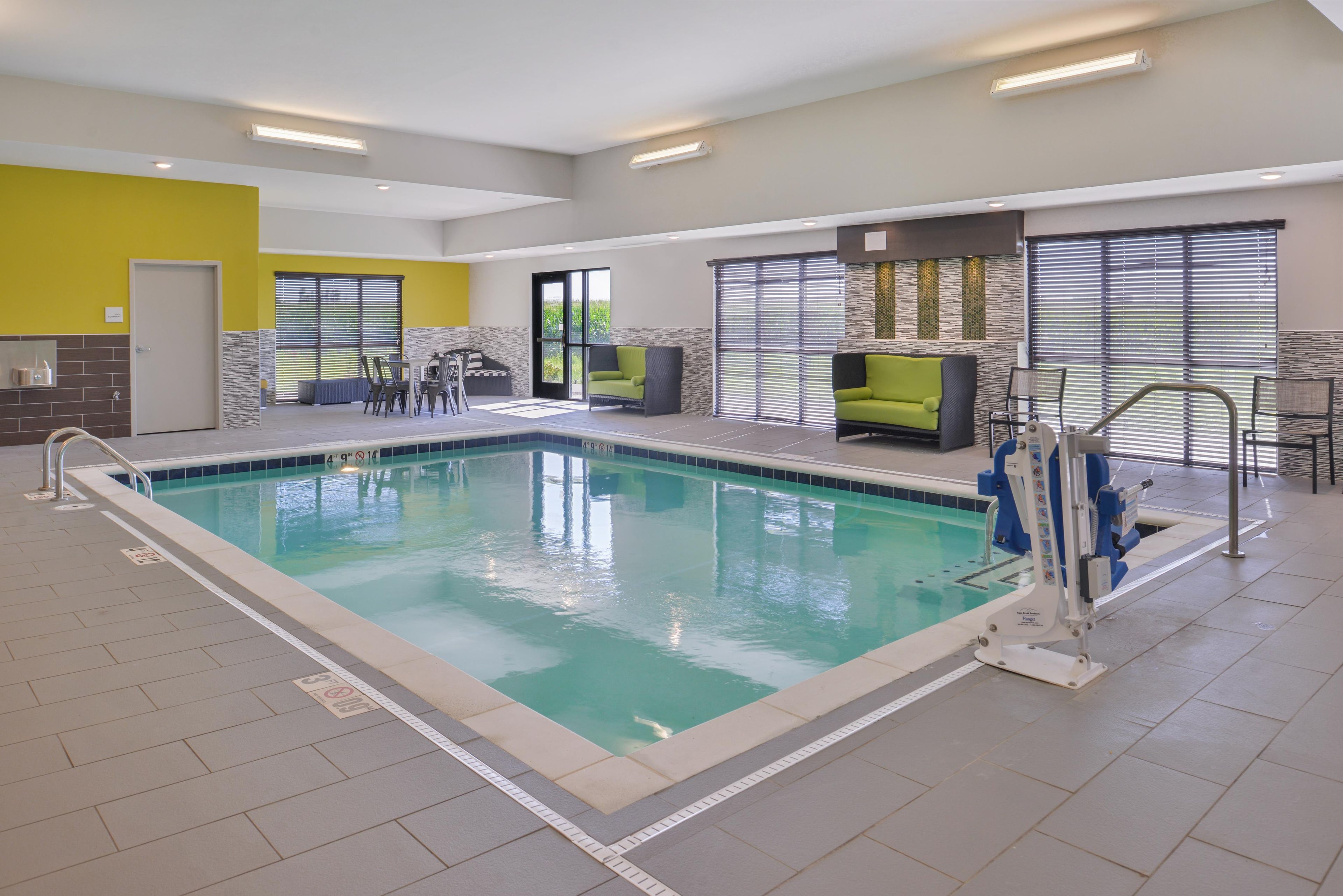 Ein Hallenbad mit Rettungsschwimmerstand, einer Sitzgelegenheit mit Stühlen und einem Tisch sowie einer Tür links.