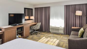 Una habitación de hotel con una cama, un escritorio con televisor, una silla, una lámpara y una ventana con cortinas.