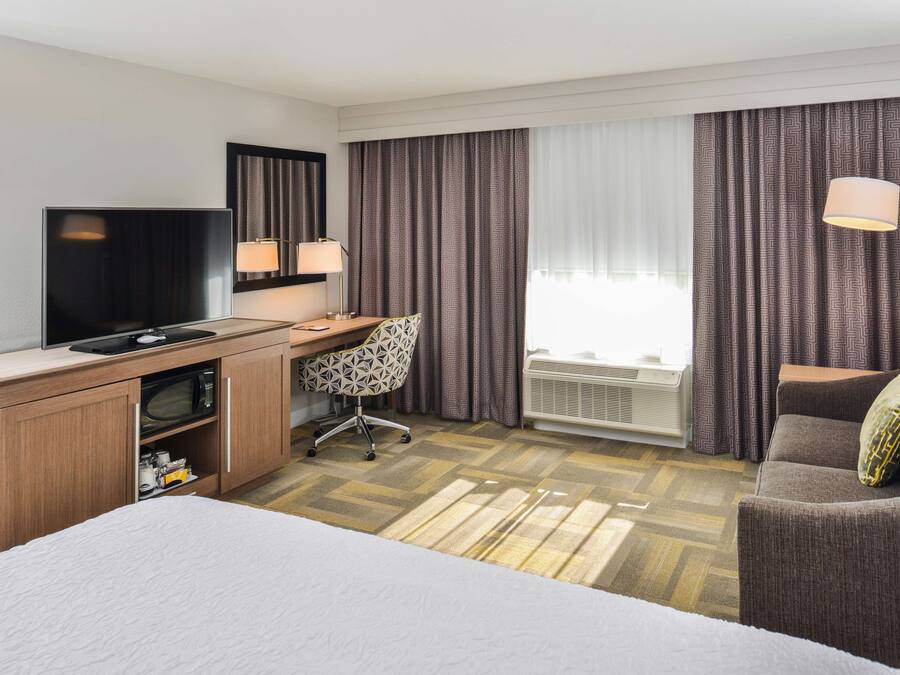 Una habitación de hotel con una cama, un escritorio con televisor, una silla, una lámpara y una ventana con cortinas.