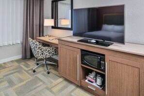 A hotel room with a bed, a flat-screen TV, a desk with a lamp, a patterned chair, and a minibar. - Hampton Inn & Suites Altoona-Des Moines (Altoona)