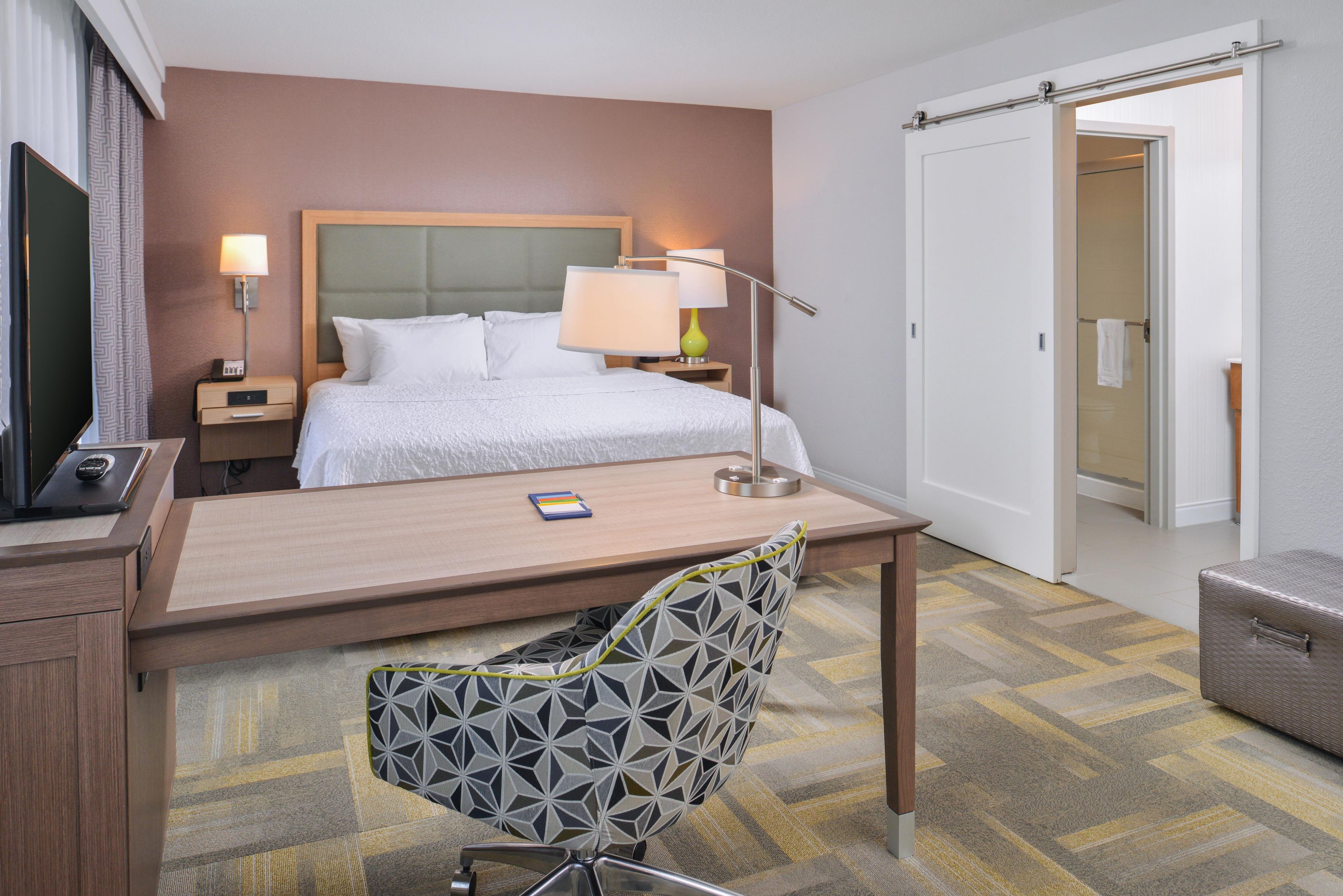 A hotel room with a bed, bedside tables, a desk, a chair, a TV, and a wardrobe.