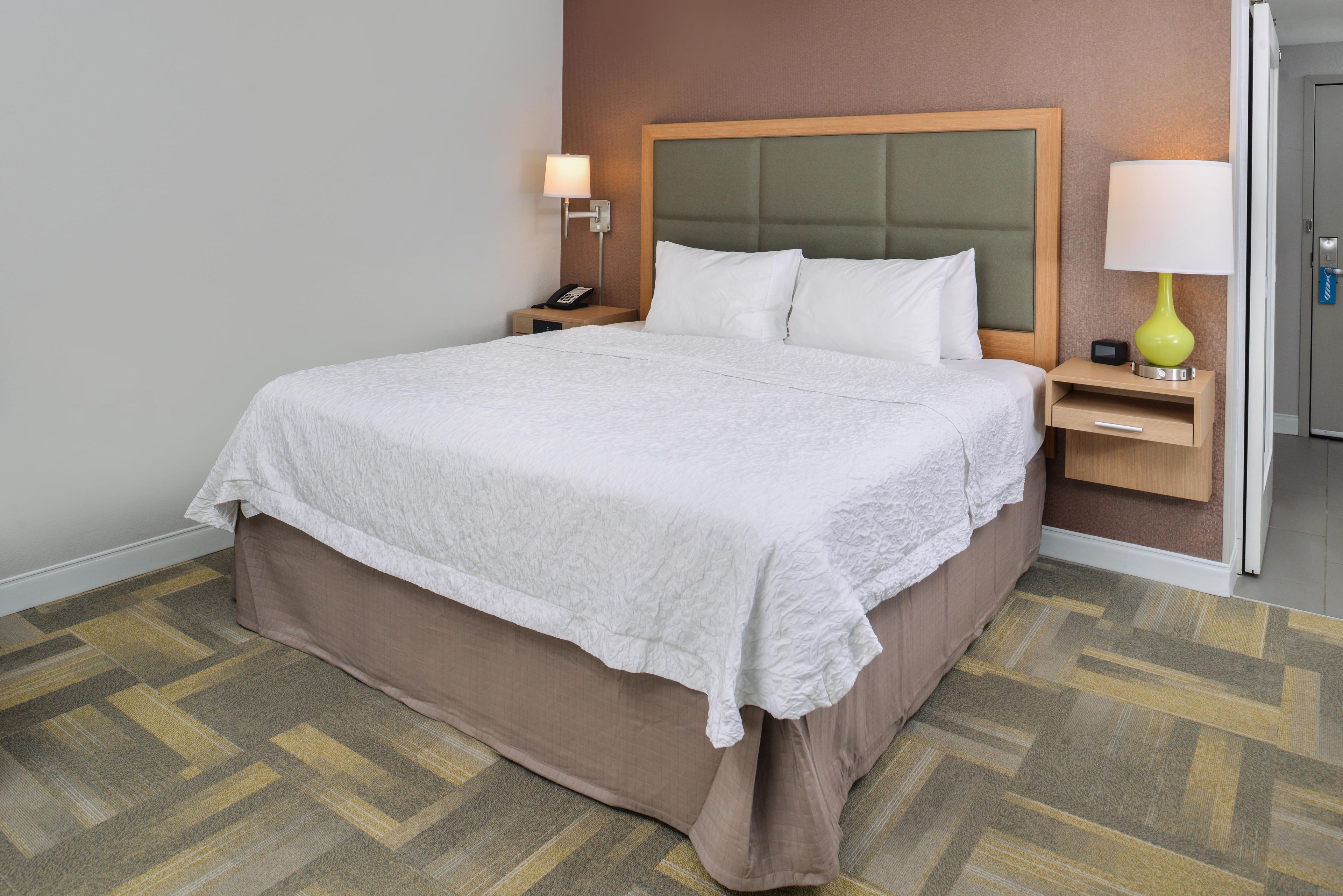 A hotel room with a large bed, a bedside table with a lamp, a phone, and a door to the right.