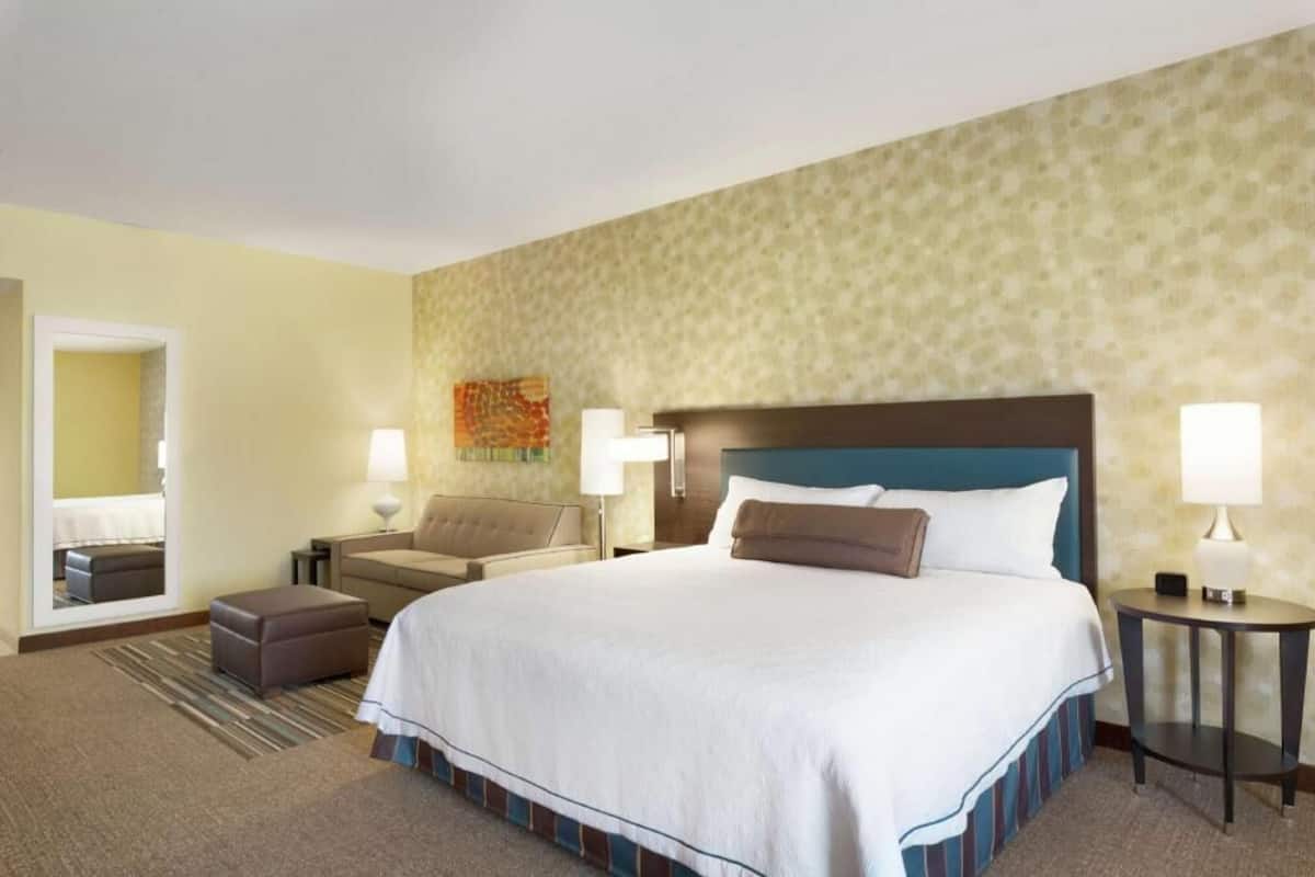 A hotel room with a large bed, two bedside tables, a sofa, a small ottoman, and a mirror.