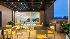 An outdoor patio area with yellow chairs and tables, a barbecue grill, and a pool in the background.