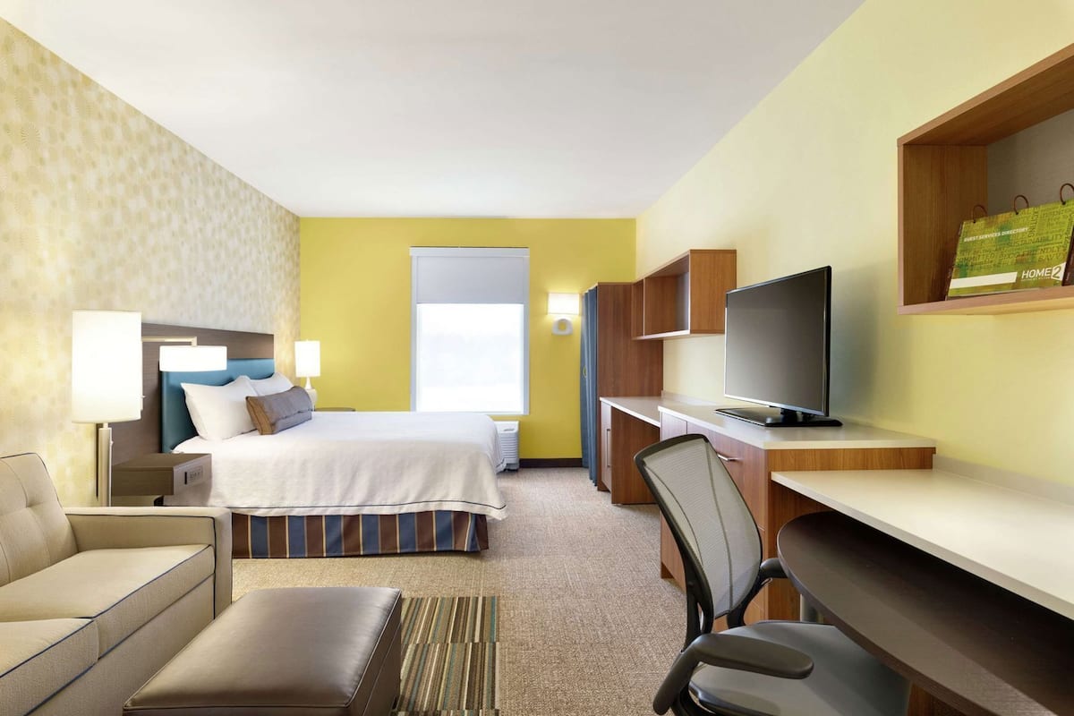 A hotel room with a bed, a desk with a computer, a TV, a chair, a sofa, and a lamp.