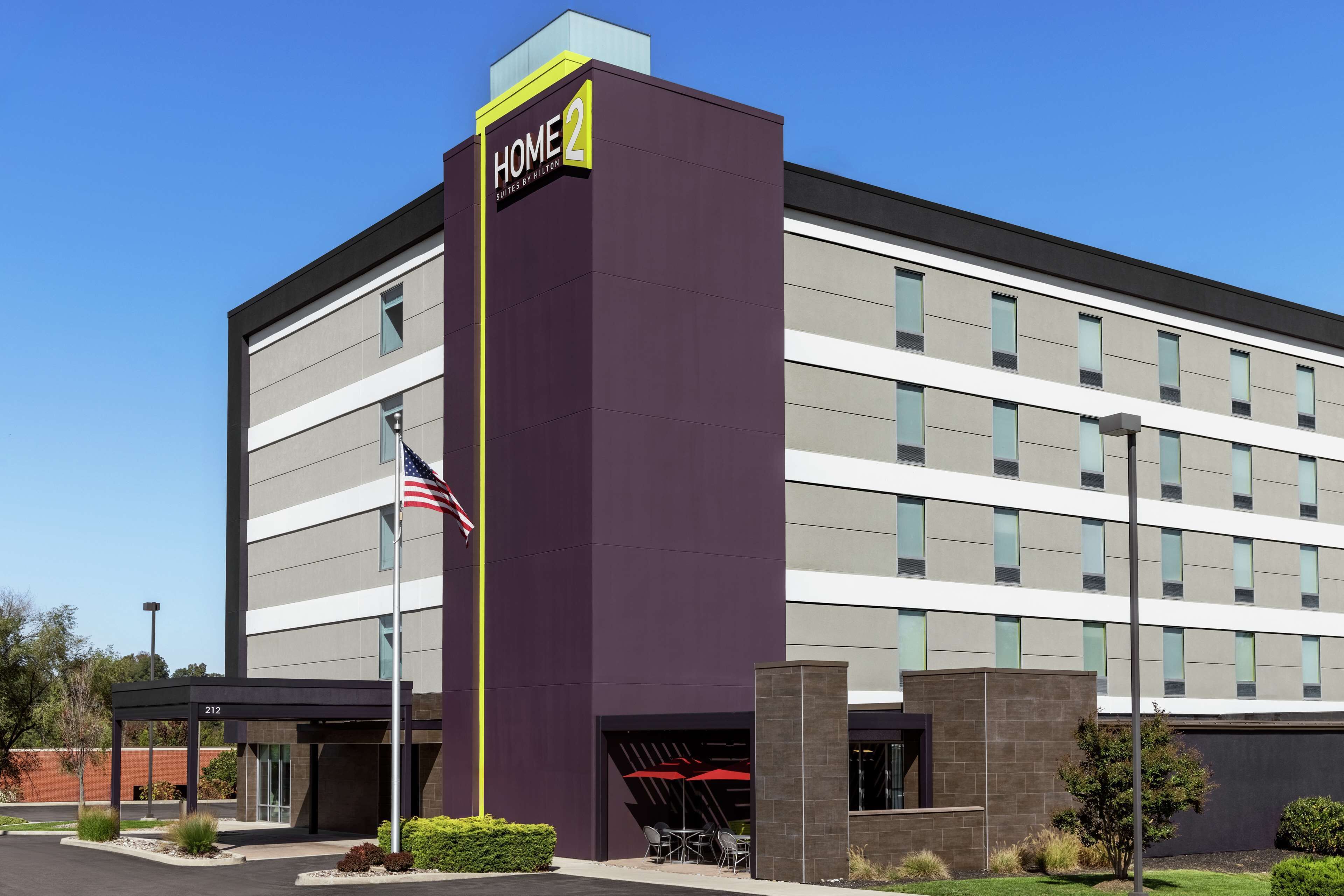 A modern hotel building with a prominent 'HOME 2' sign, a flagpole with an American flag, and a covered outdoor seating area.