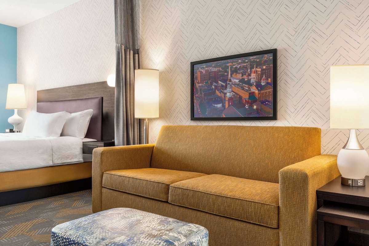 A hotel room with a bed, a sofa, a coffee table, and a cityscape painting on the wall.