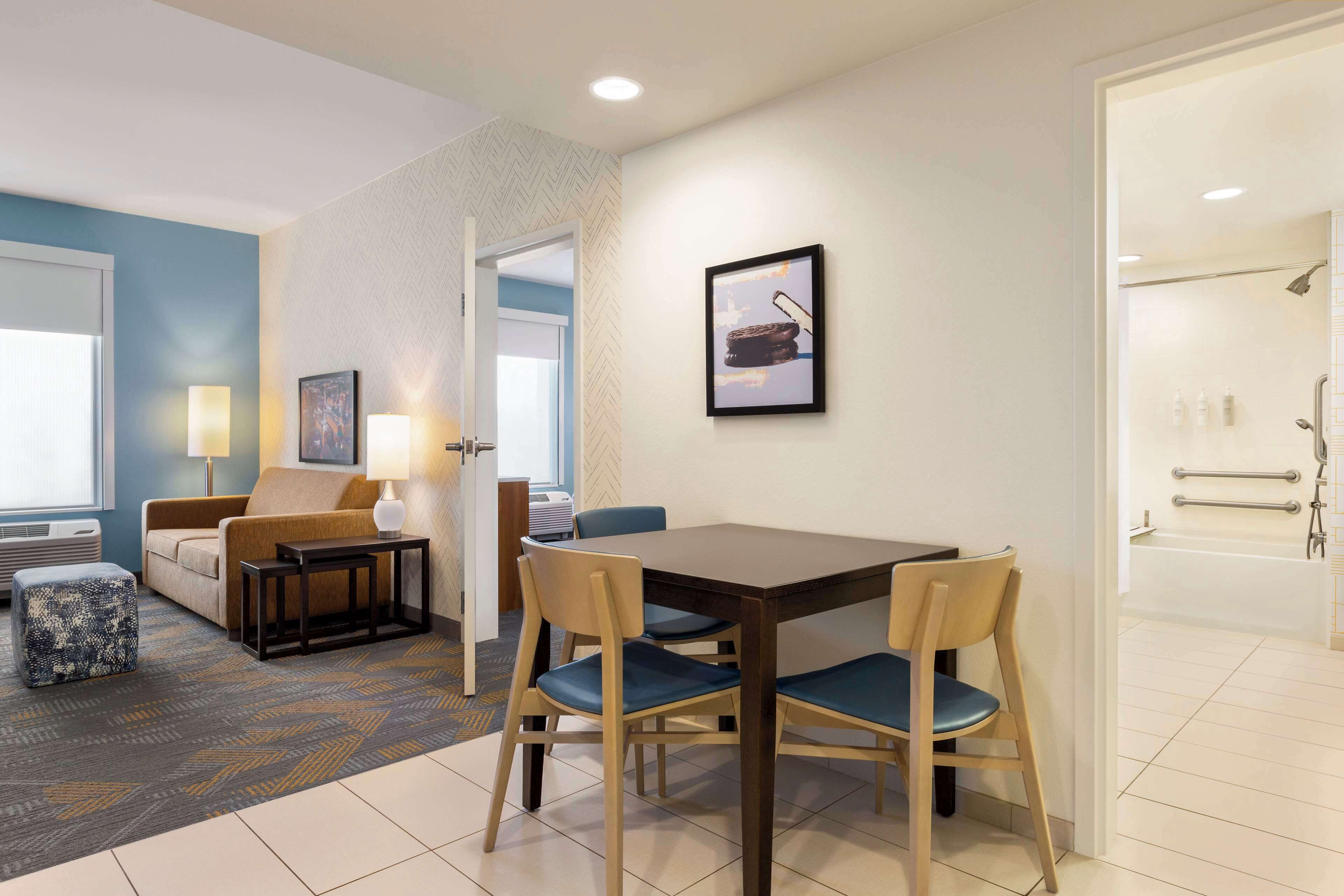 A hotel room with a dining area, a sofa, a coffee table, and a bathroom with a bathtub and shower.