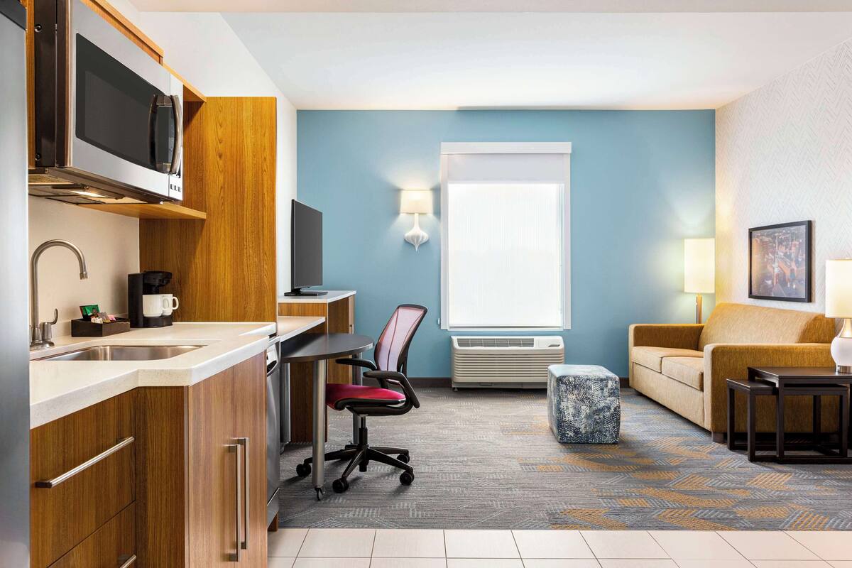 A compact hotel room with a kitchenette, a dining area, a seating area with a sofa, and a large window providing natural light.