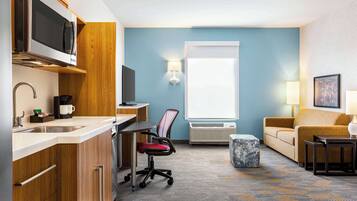 A compact hotel room with a kitchenette, a dining area, a seating area with a sofa, and a large window providing natural light.