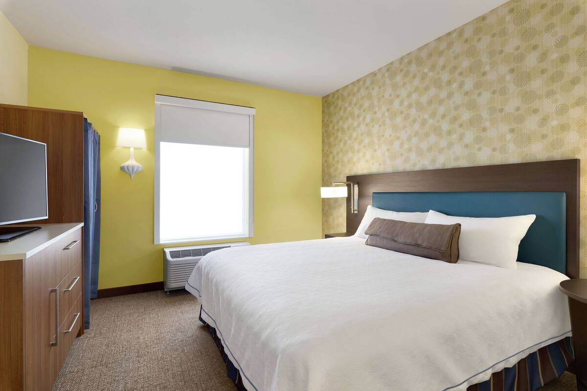 A hotel room with a bed, a desk with a television, a window with blinds, and a patterned wall.