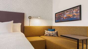 Habitación de hotel con cama, sofá, una mesita, lámpara y un cuadro con vista a la ciudad.