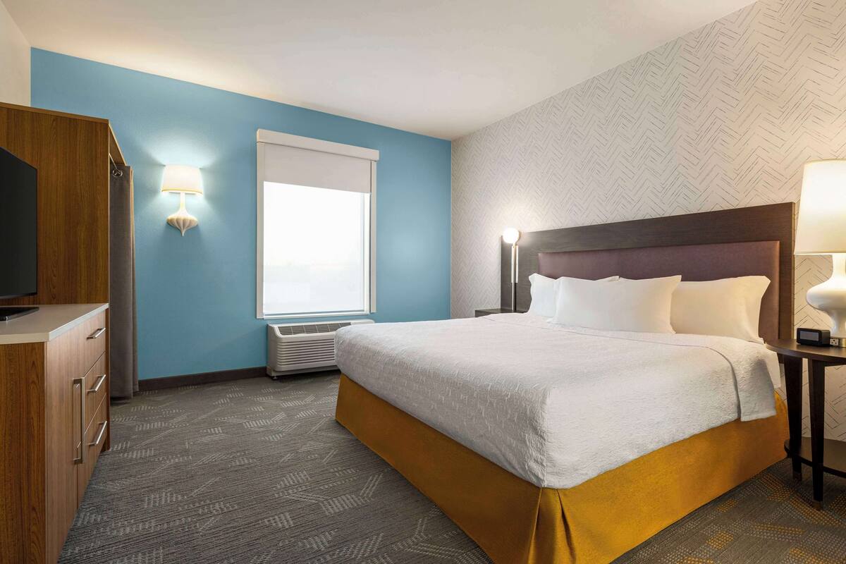 A hotel room with a bed, a TV, a desk, and a window with blinds.