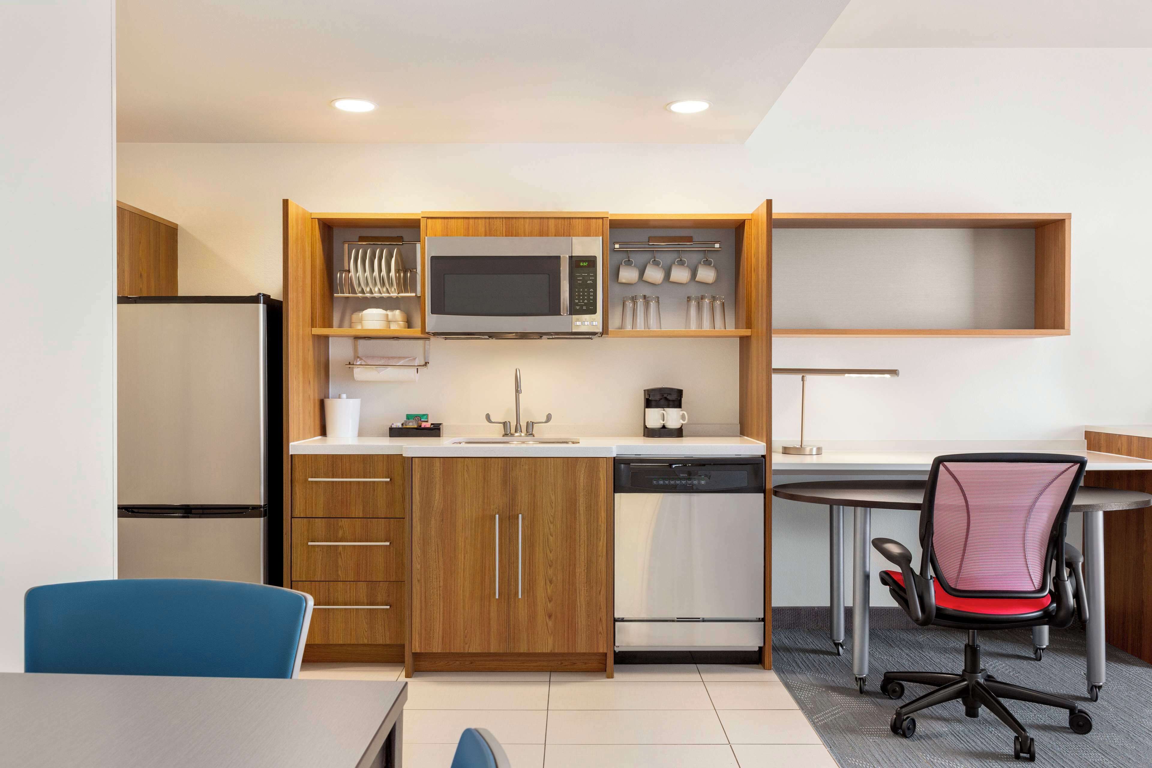 A compact kitchen with a microwave, coffee maker, and dishwasher. There is a dining table with a blue chair and a red office chair.
