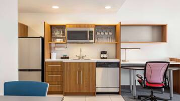 A compact kitchen with a microwave, coffee maker, and dishwasher. There is a dining table with a blue chair and a red office chair.
